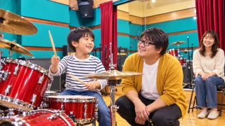 ピクミン講師と小学生が笑顔でドラムを練習し保護者が見守る様子